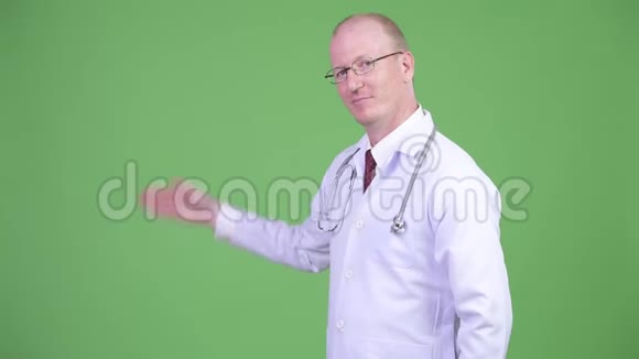 快乐成熟的秃头医生在绿色背景下展示一些东西视频的预览图