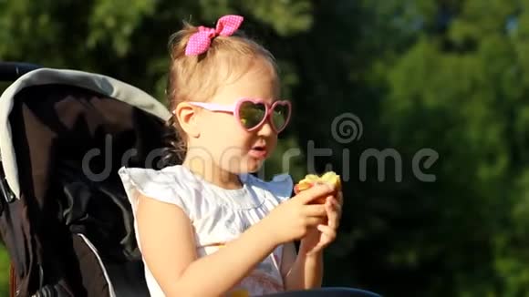 戴太阳镜的孩子坐在婴儿车里日落时吃苹果肖像女婴特写在阳光明媚的日子视频的预览图