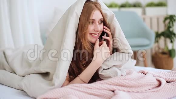 躺在毯子下躺在床上用手机聊天的年轻红发女孩视频的预览图