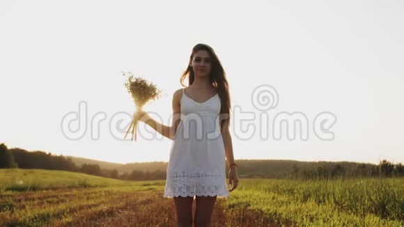 穿着白色裙子的女人站在田野里拿着一束鲜花看着镜头微笑着视频的预览图
