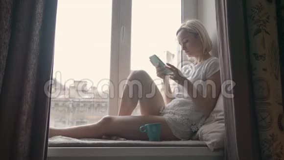 年轻女子在家中坐在窗台上喝咖啡使用智能手机视频的预览图