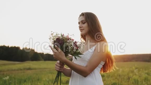 快乐的年轻女子穿着白色连衣裙站在田野里夕阳下捧着一束鲜花视频的预览图