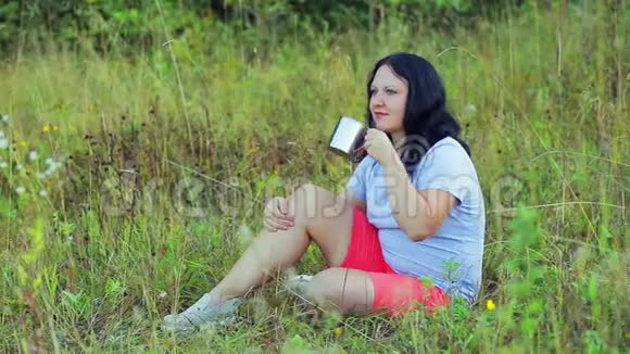 年轻快乐的女人坐在草地上喝茶欣赏大自然视频的预览图