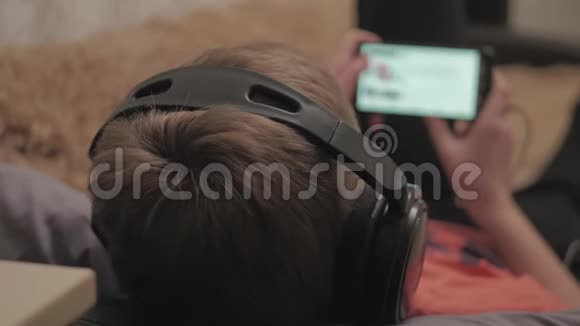 男孩看媒体内容使用耳机和手机应用程序躺在家里的沙发上把男孩的手收起来视频的预览图