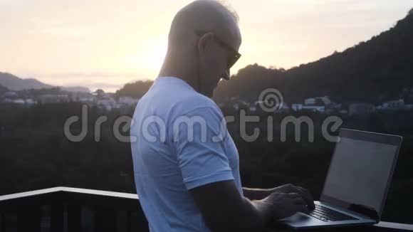 一位英俊的年轻人站在美丽的日出的屋顶上在笔记本电脑上工作视频的预览图