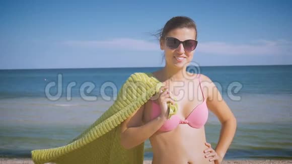 穿着泳装和太阳镜的女孩微笑着站在海边在海风中拿着海滩毛巾蓝色绿色视频的预览图