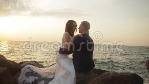 幸福迷人的新婚夫妇正坐在岩石上温柔地拥抱着在大海的背景下交谈着视频的预览图