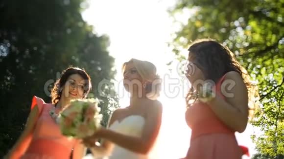 迷人的金发新娘正在阳光明媚的公园里向空中投掷婚礼花束她和她的两个人在一起视频的预览图