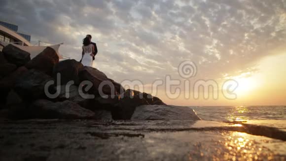 日落时岩石上的波浪破碎美丽的一对新婚夫妇站在上面时温柔地拥抱着视频的预览图