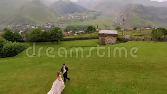 美丽而快乐的新郎新娘在雄伟的群山背景下沿着绿草奔跑视频的预览图
