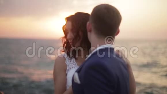 可爱的新娘和英俊的新郎温柔地拥抱在海边的背景下明亮的五颜六色的日落视频的预览图