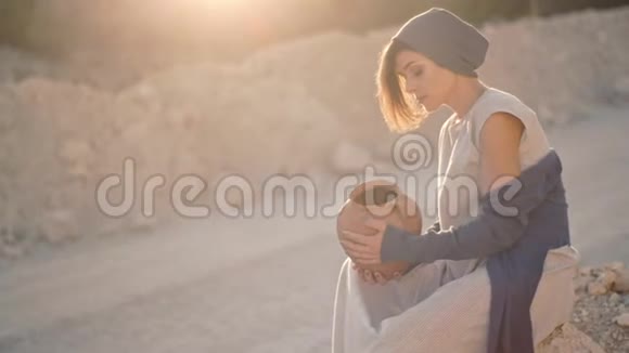 孤独悲伤的女人坐在沙路和石头的背景上有一个空的粘土花瓶视频的预览图