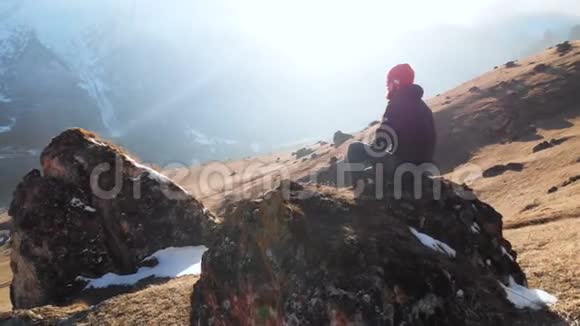 一个戴太阳镜的留胡子的嬉皮士正坐在山坡上的一块大石头上背景是白雪皑皑视频的预览图