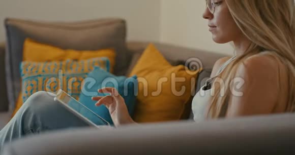 高质量的4k12位镜头美丽的女孩使用平板电脑坐在家里的沙发上女人上网社交视频的预览图