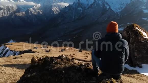 一个戴太阳镜的留胡子的嬉皮士正坐在山坡上的一块大石头上背景是白雪皑皑视频的预览图