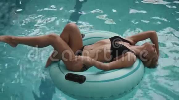 迷人的年轻金发瘦女人穿着黑色比基尼躺在游泳池里的充气橡胶圈上4k视频的预览图