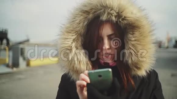 使用智能手机技术应用程序的美女穿着一件带绒毛罩的保暖夹克在街上行走的特写镜头视频的预览图