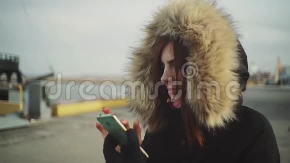 使用智能手机技术应用程序的美女穿着一件带绒毛罩的保暖夹克在街上行走的特写镜头视频的预览图