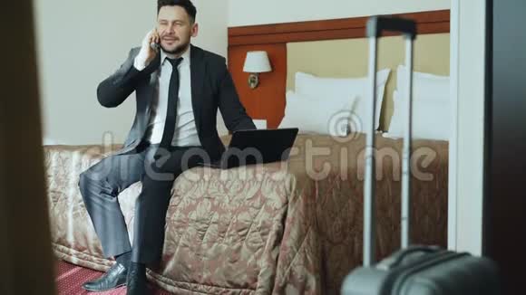 穿着西装穿着笔记本电脑坐在酒店房间的床上用手机说话的微笑商人的倾斜视频的预览图