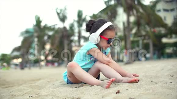 海滩上的小女孩戴着太阳镜戴着白色耳机听音乐在海滩上玩沙子视频的预览图