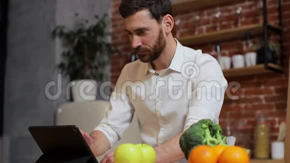 男人在家庭厨房的平板电脑上浏览英俊的年轻人在平板电脑上浏览微笑着快乐一个男人吃视频的预览图