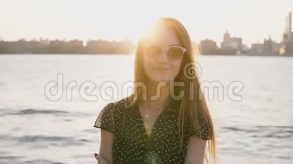 年轻美丽的调情欧洲女孩的肖像戴着太阳镜对着镜头微笑在纽约市河流日落4K视频的预览图