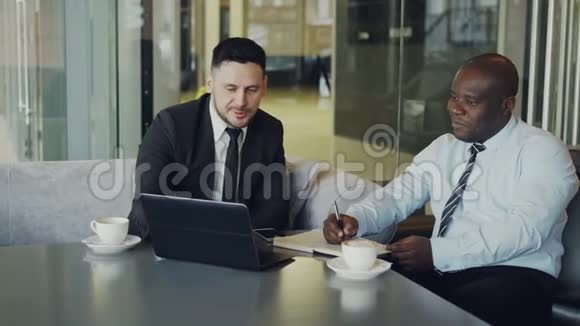 两个兴高采烈的商人在现代咖啡馆讨论项目非裔美国人的企业家写笔记本和他的视频的预览图