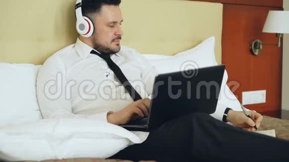 快乐的商人戴着耳机在笔记本电脑上工作在酒店房间里躺在床上时微笑着听音乐视频的预览图