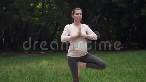 孕妇在公园里做瑜伽的地毯上站在一条腿上的姿势体式树式视频的预览图