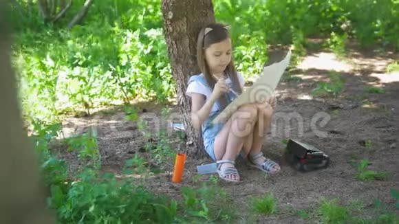 夏天白种人的小女孩坐在树下用画笔画画视频的预览图