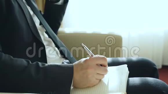 一位自信的商人坐在扶手椅上时会在笔记本上用手提电话和书写笔记视频的预览图