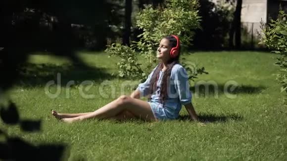 在阳光明媚的天气里小女孩坐在公园的草地上听耳机上的音乐4K视频的预览图