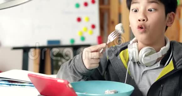 年轻的亚洲男孩在家里用耳机在平板电脑上吃和玩多莉向左向右移动视频的预览图