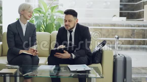 两位女性和男性商务同事在酒店大堂使用数字平板电脑记事本和讨论项目视频的预览图