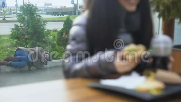 无家可归的人躺在街上而快乐的年轻女子在咖啡馆里吃汉堡视频的预览图