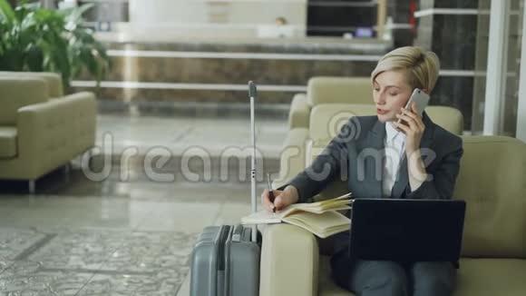 潘在酒店大厅里坐在扶手椅上的金发忙碌的女商人用手提电话记事本和视频的预览图