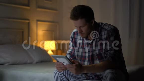 不快乐孤独的年轻男性坐在床上看照片分手思念妻子视频的预览图