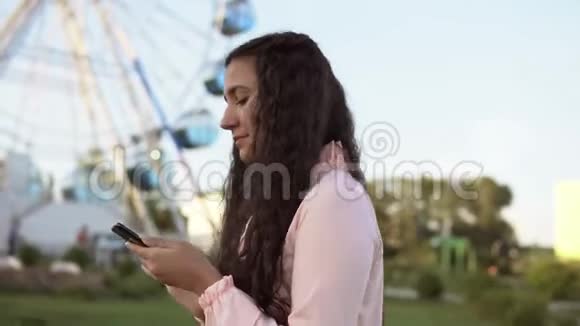 一个年轻的女孩走在摩天轮旁使用智能手机4K视频的预览图