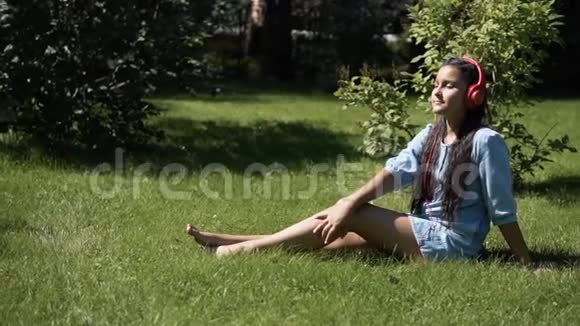 戴着深色长发的小女孩坐在公园的草地上听着耳机里的音乐视频的预览图