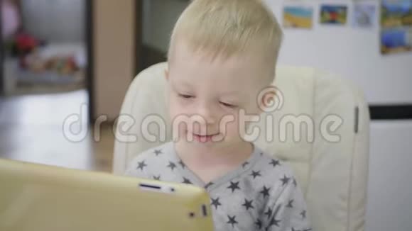 可爱的小男孩在家吃粥吃早餐同时在平板上看卡通儿童和技术视频的预览图