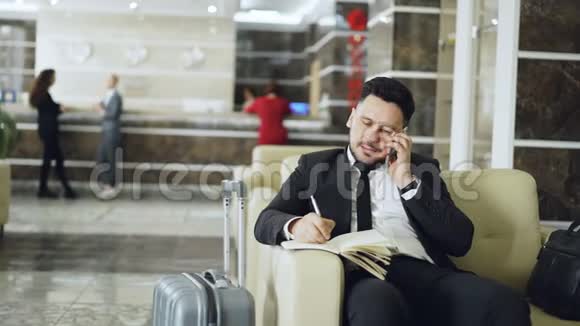 潘在坐在豪华的扶手椅上时拍了一张自信的商人在谈论手机和写笔记的照片视频的预览图