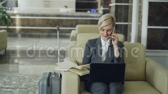 潘在酒店大厅里坐在扶手椅上的金发忙碌的女商人用手提电话记事本和视频的预览图
