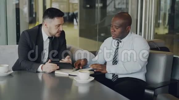 两个穿着正式服装的多民族伙伴看着数码平板电脑在餐桌上讨论他们的商业想法视频的预览图