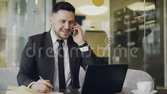 身穿黑色商务服满脸胡子的快乐商人坐在餐桌前微笑着用现代手机交谈视频的预览图