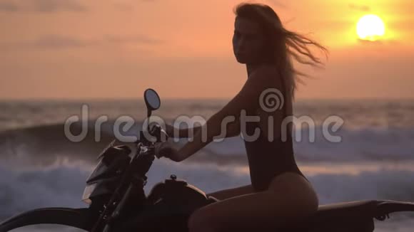 可爱的金发女郎坐在赛车摩托车上红色的夕阳和傍晚暴风雨的大海在她身后视频的预览图