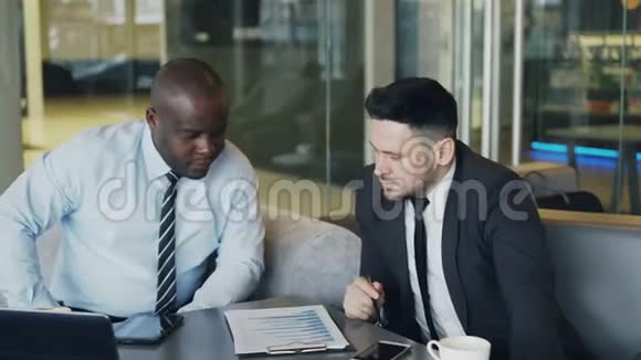 两个穿着正式服装的多民族伙伴坐在一起一边看着记事本一边讨论财务图表的细节视频的预览图