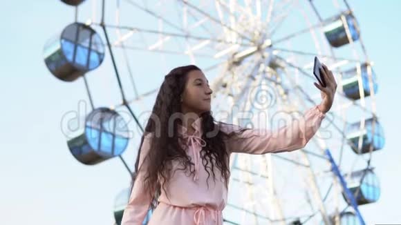 一个穿着粉红色长裙的长发女孩站在摩天轮附近用手机自拍慢慢慢慢视频的预览图