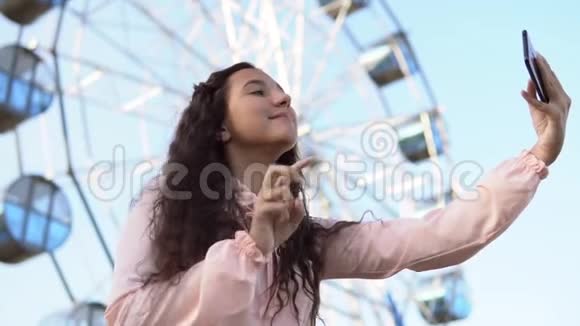 一个穿着粉红色连衣裙的长发女孩用站在摩天轮附近的手机自拍慢动作视频的预览图