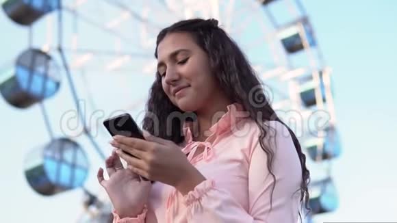 女孩用智能手机站在大摩天轮和游乐园附近慢动作视频的预览图