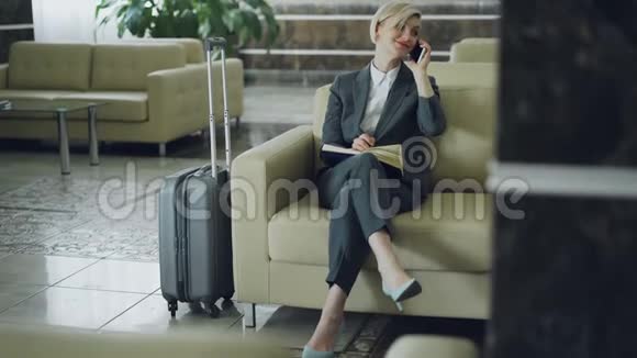 漂亮的女商人坐在酒店大堂的扶手椅上用手机说话用记事本写字微笑着视频的预览图